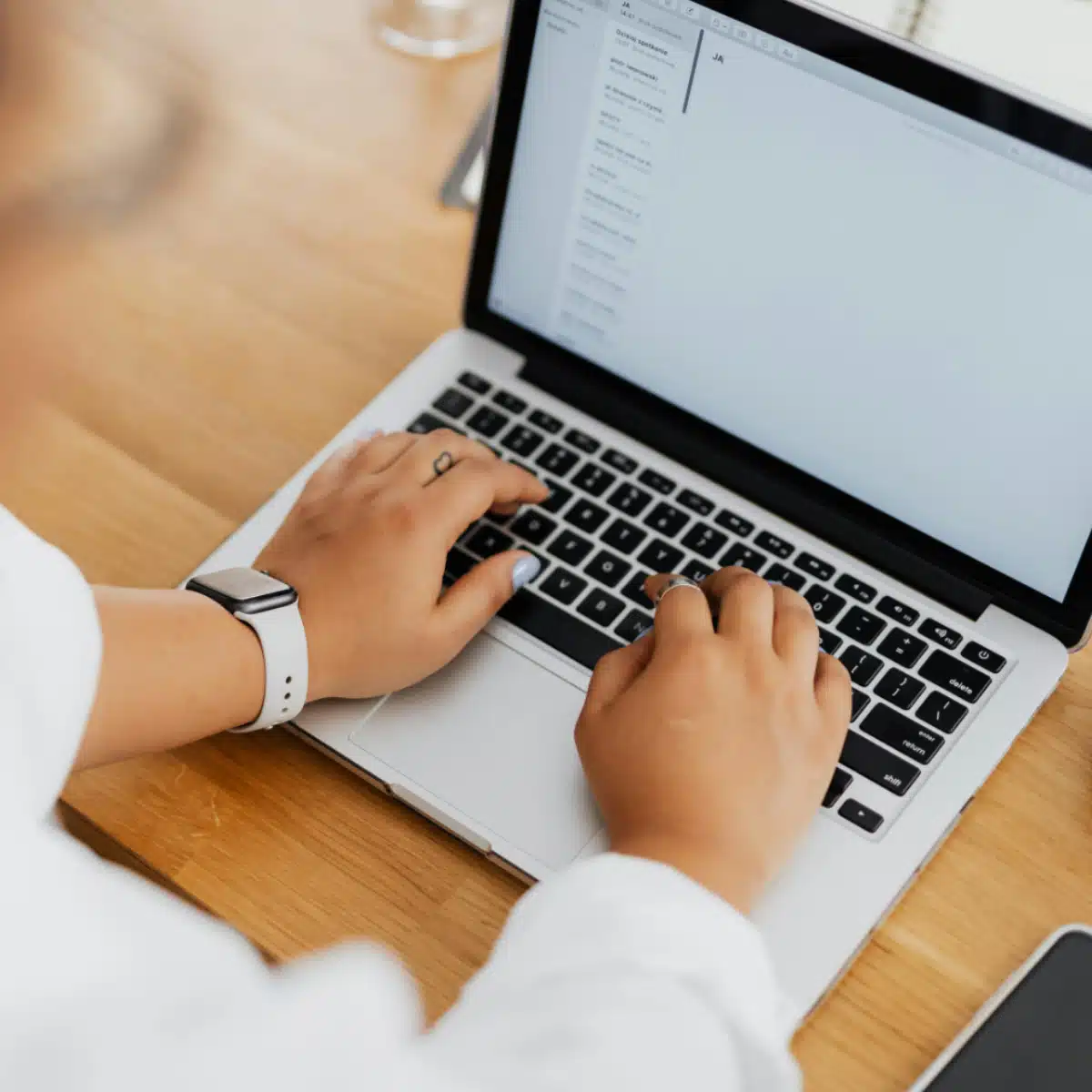 Person wearing a smartwatch typing on a laptop at a wooden desk, with an email or message inbox open on the computer screen.