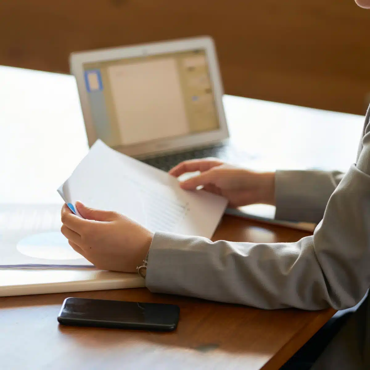 A person in a gray suit reviews printed documents with charts at a wooden desk. An open laptop and a smartphone are also on the desk. The person's face is not visible.