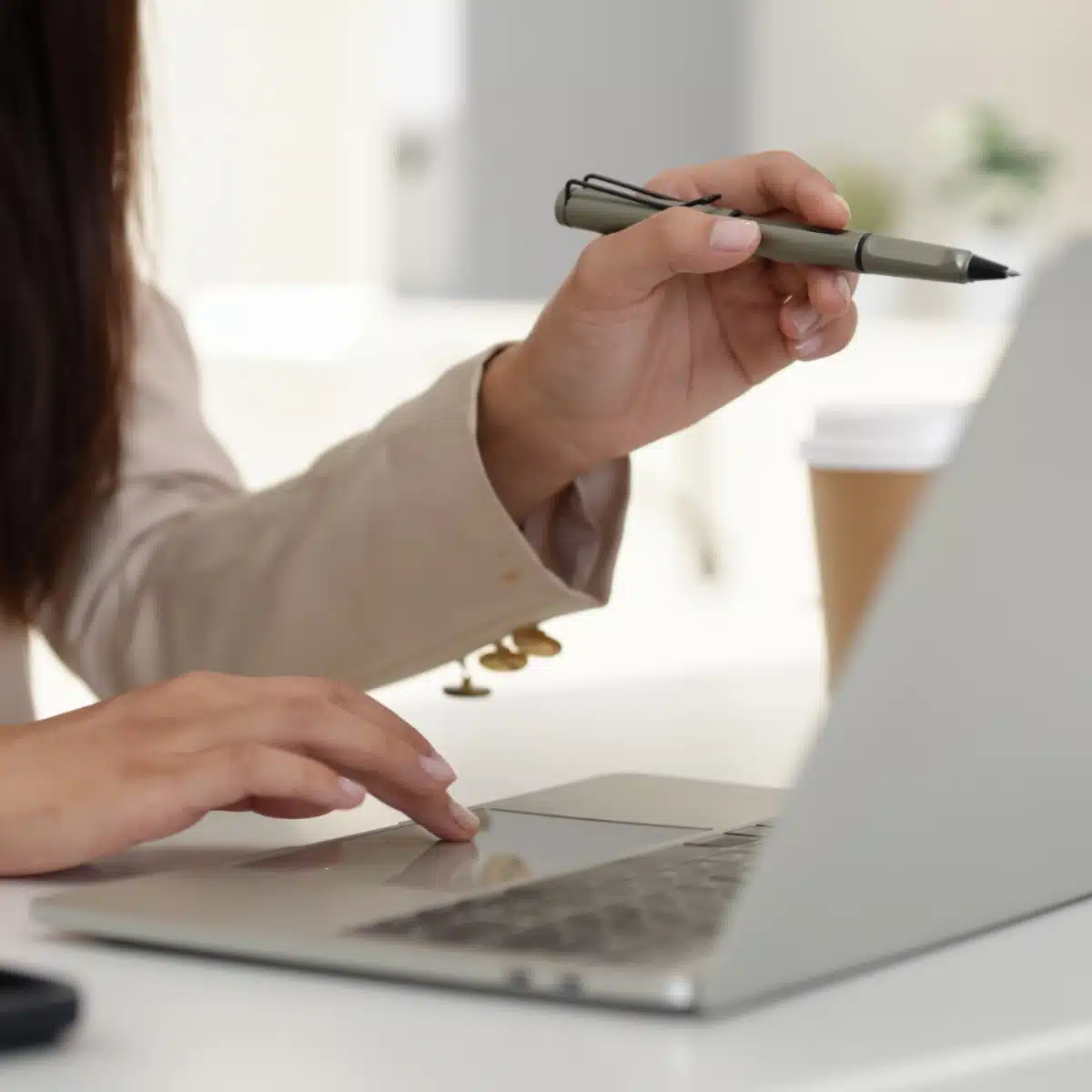 A person holding a pen and using a laptop at a desk, with a takeaway coffee cup in the background. Only their hands and part of their arm are visible.