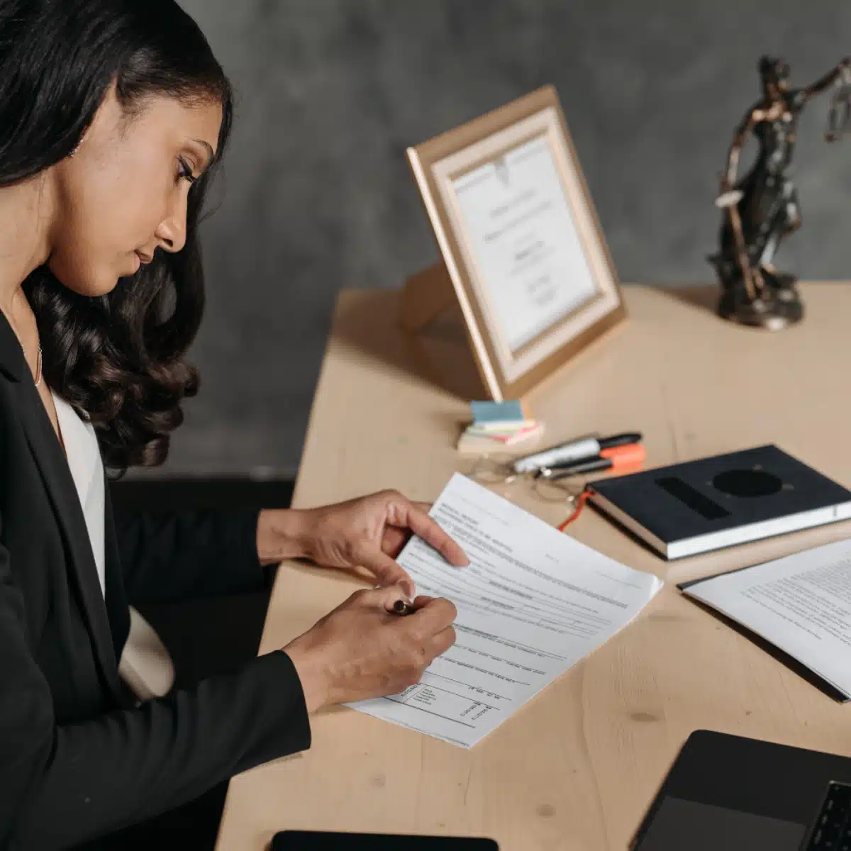 A woman in business attire sits at a desk, focused on signing documents. The desk has pens, sticky notes, a closed notebook, and a framed certificate in the background, with a statue of Lady Justice beside it.