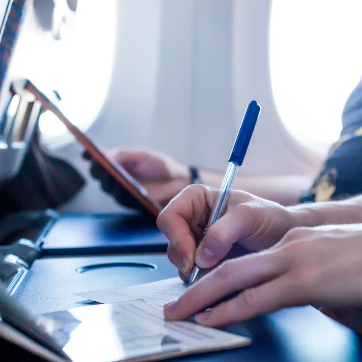 A person writes on a piece of paper with a blue pen on an airplane tray table, while another person uses a tablet nearby. Bright daylight is visible through the airplane windows in the background.