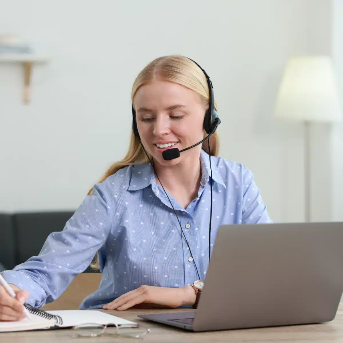 A woman wearing a headset smiles while working at a desk with a laptop, writing in a notebook. She appears to be engaged in a video call or virtual meeting in a home office setting.
