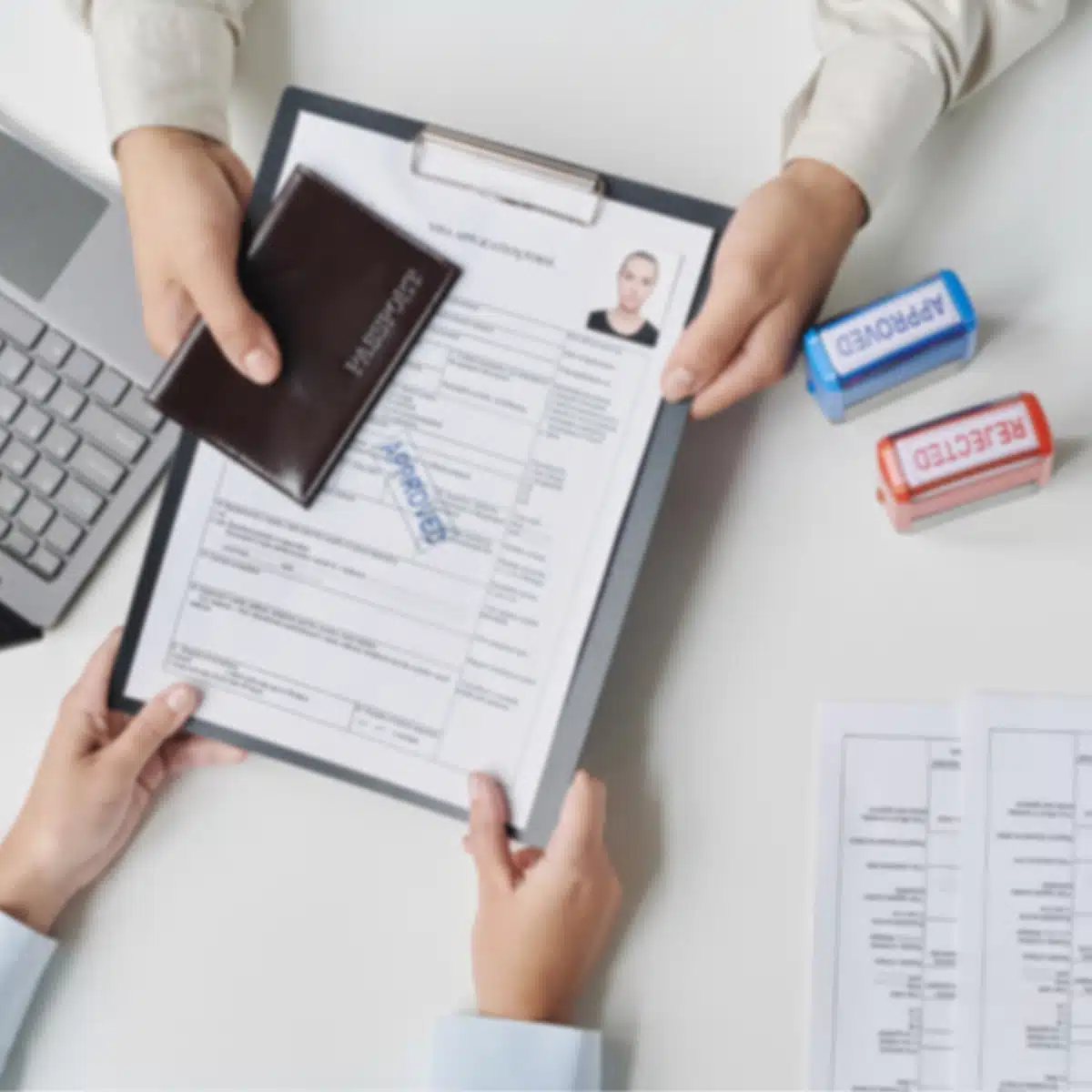 A person hands over a clipboard with a job application form and passport, while another person reviews it. "Rejected" and "Approved" stamps are nearby, along with a laptop and additional documents on the desk.