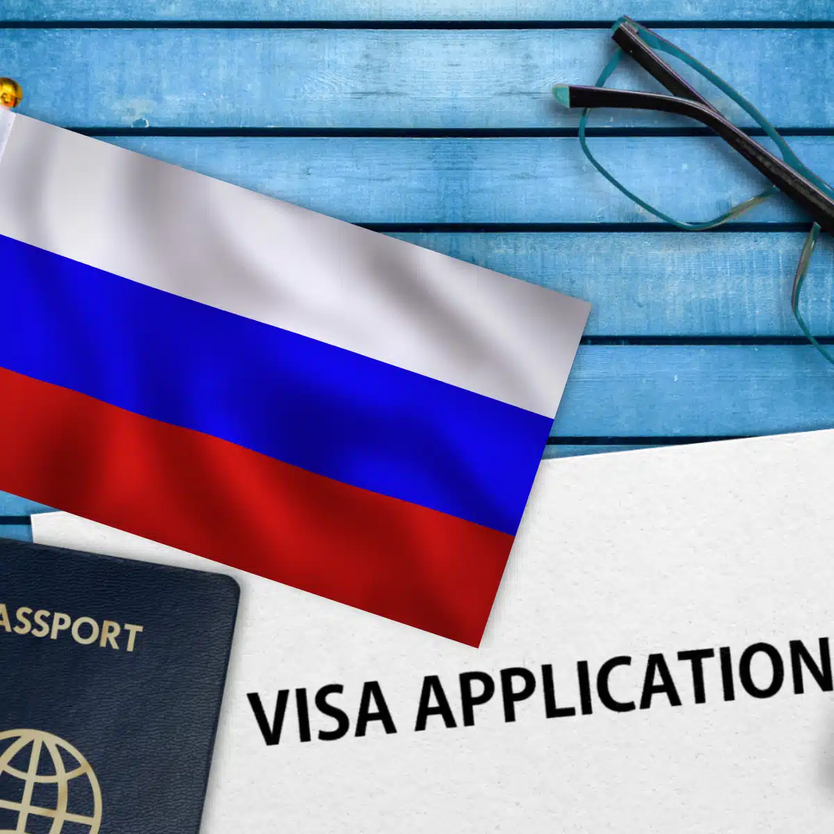 A Russian flag, a pair of glasses, a passport, and a document labeled "VISA APPLICATION" are arranged on a blue wooden surface.